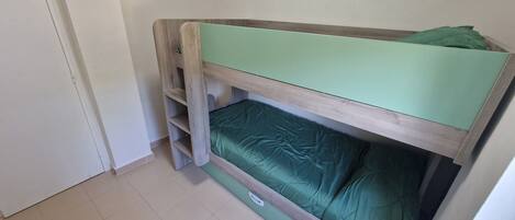 Una cama litera con colchón verde y estructura de madera en una habitación con paredes blancas y suelo de baldosas.
