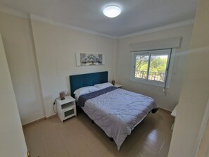 Een slaapkamer met een bed, nachtkastje, wandplank, raam met uitzicht en een plafondlamp.