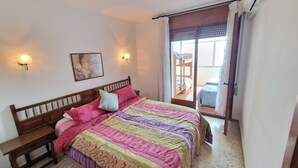 A bedroom with a wooden bed, a bench, a wall-mounted light, a painting, and a sliding door leading to a balcony.