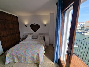 A bedroom with a bed, a heart-shaped wall decoration, a window with a view of boats, and a balcony. - VILLA EMPORION CANALS (Empuriabrava)