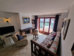A living room with a TV, sofa, armchair, fireplace, and a view of a water body through a glass door. - VILLA EMPORION CANALS (Empuriabrava)