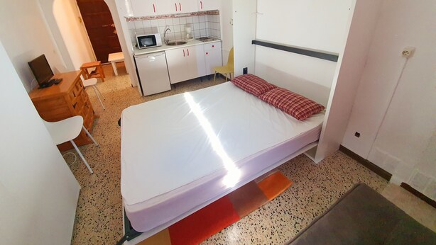 Ein kompakter Raum mit Bett, Schreibtisch, Stuhl, einer kleinen Küchenzeile und einer Bank.