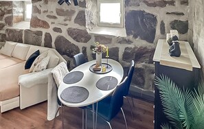 A stone-walled room with a beige sofa, a dining table with four chairs, and a small plant.