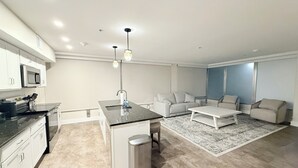 Une cuisine moderne avec des meubles blancs, un plan de travail noir et un évier en acier inoxydable. On y trouve un coin repas avec un canapé blanc et une table basse. La pièce est décorée dans des tons neutres et possède une grande fenêtre.