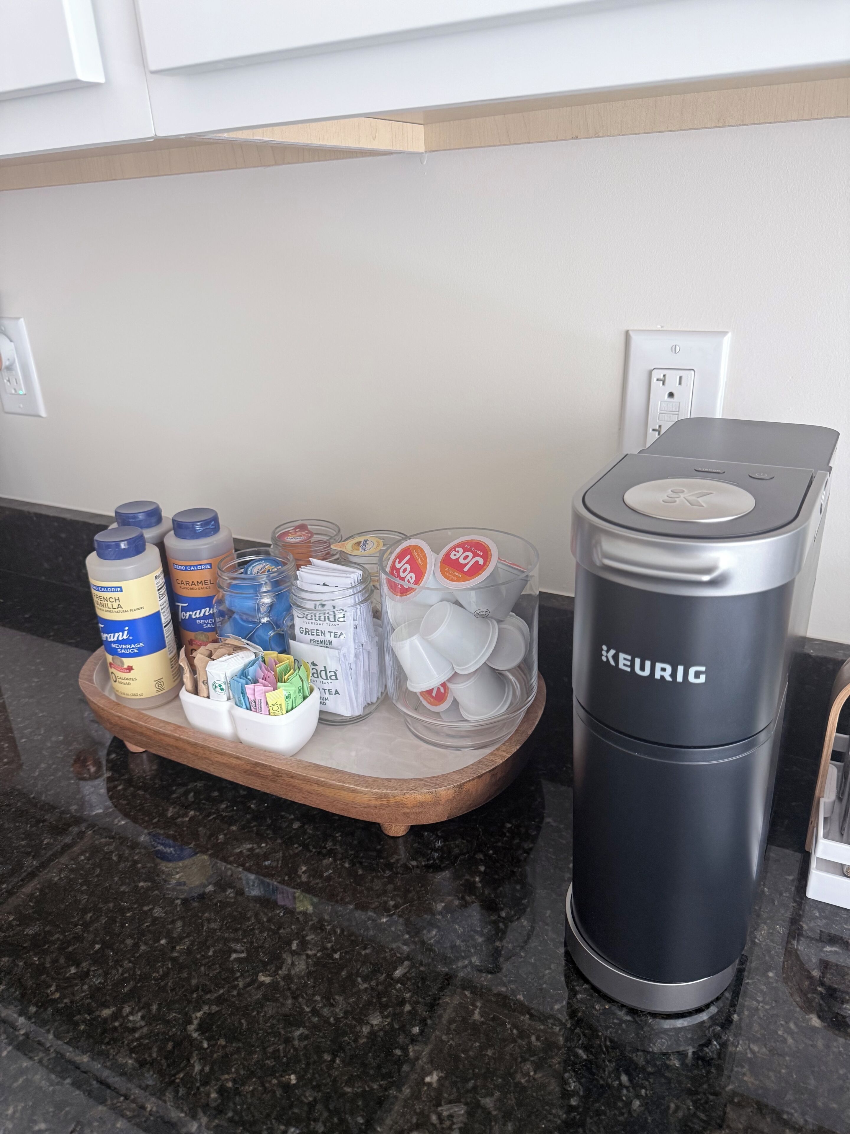 A kitchen counter with a Keurig coffee maker, a tray with various coffee pods, and a container of creamer.