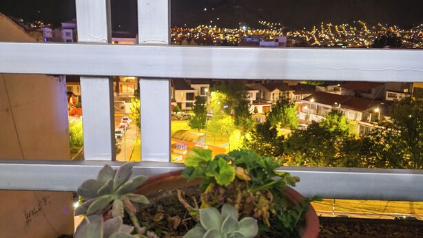 A balcony with a potted plant, a railing, and a view of a cityscape at night. - Upscale 3BR Apt in Cusco,Gateway to Machu Picchu , Best Area (Cusco)