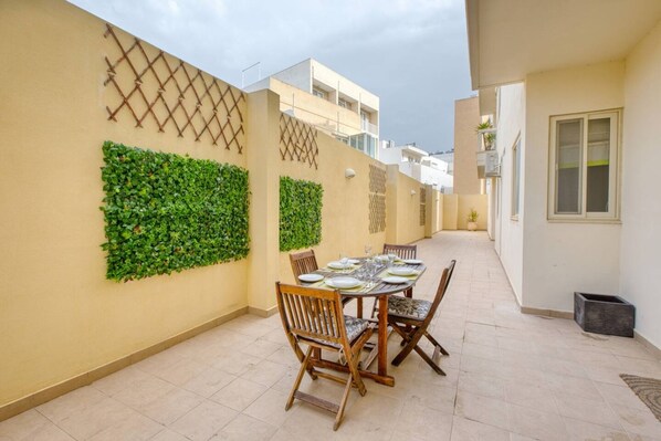 Uma área de refeições com mesa posta para quatro pessoas, rodeada de cadeiras, parede decorativa e vista desobstruída para a rua.