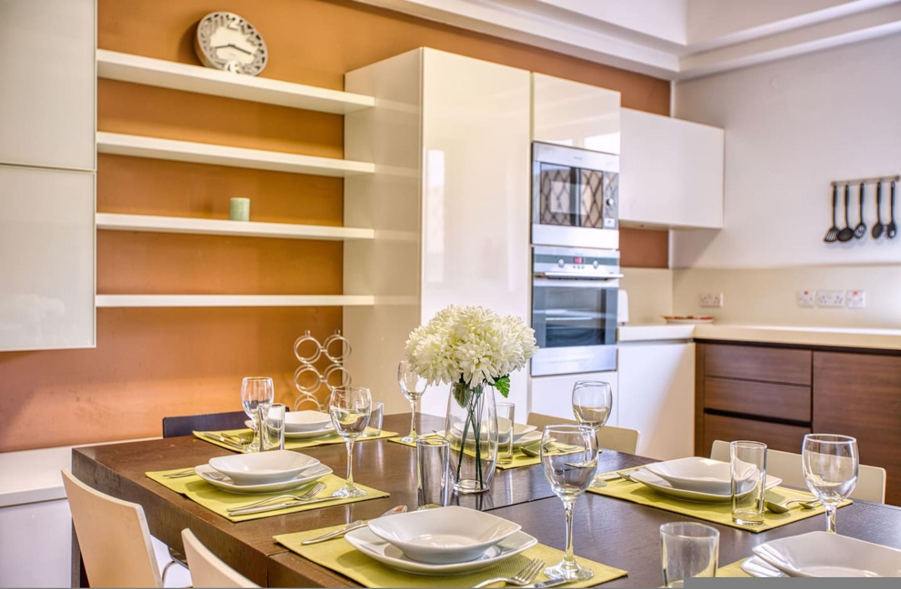A modern kitchen with white cabinets, a built-in oven, and a dining area set with white plates and glasses.