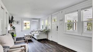 A spacious room with a large window, a sofa, a chair, and a small table. - Saratoga Lake, 3 Cottages + Private Beach With Dock (Saratoga Springs)