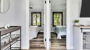 A room with a bed, a white wardrobe, a shelf with glassware, and a door leading to another room. - Saratoga Lake, 3 Cottages + Private Beach With Dock (Saratoga Springs)