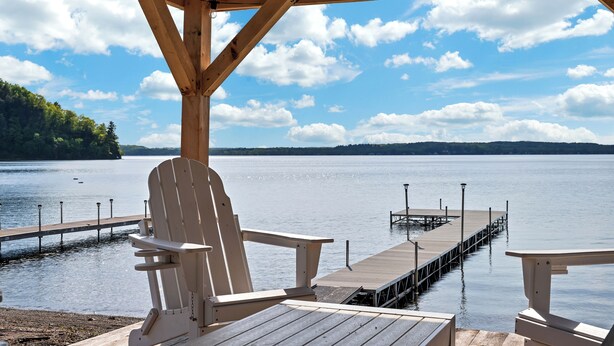 A wooden deck with Adirondack chairs overlooking a calm lake, a dock extending into the water, and a clear sky with clouds. - Saratoga Lake, 3 Cottages + Private Beach With Dock (Saratoga Springs)