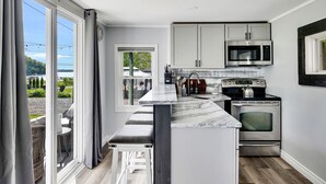 A modern kitchen with a large island, stainless steel appliances, and a view of a garden through the sliding glass doors. - Saratoga Lake, 3 Cottages + Private Beach With Dock (Saratoga Springs)