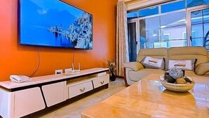 Ein Wohnzimmer mit großem Flachbildfernseher an der Wand, einer Couch, einem Couchtisch und einem Fenster mit Vorhängen.