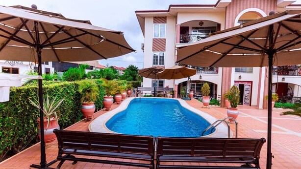 Una piscina con dos bancos, rodeada de sombrillas y un edificio con balcones al fondo.