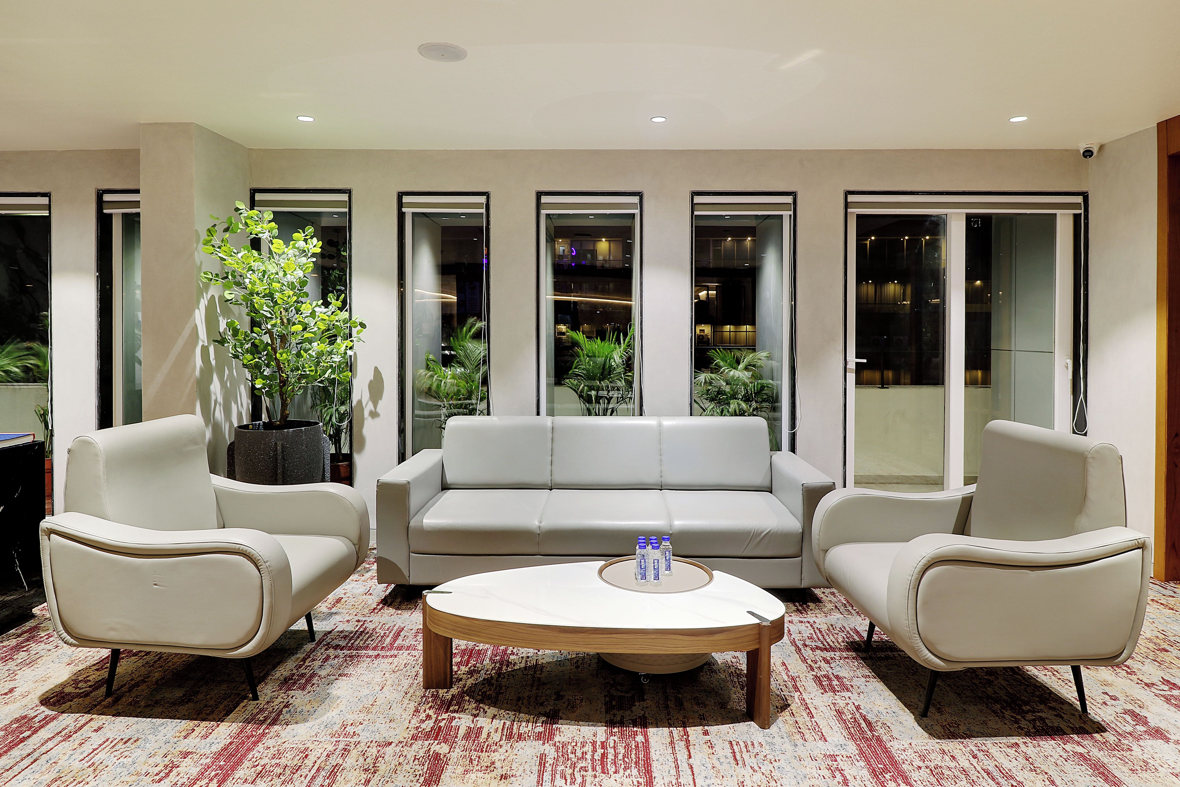 Un salón moderno con un sofá blanco, dos sillones, una mesa de centro y un ventanal con vistas a plantas de interior.