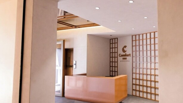 Tauleller de recepció amb acabat de fusta, un cartell fixat a la paret per 'Comfort Hotel' i un panell japonès tradicional amb un disseny de reixeta.