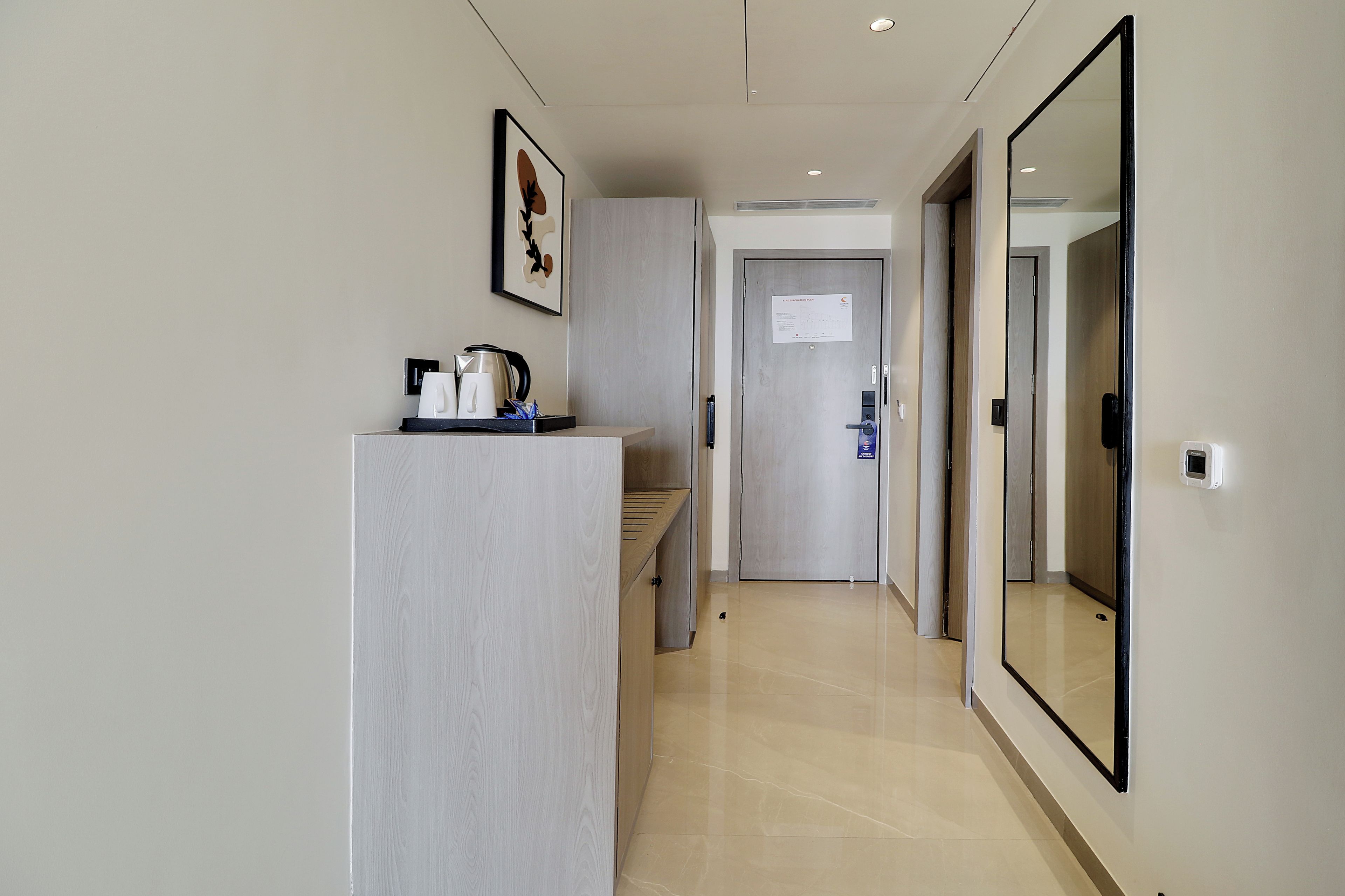 A hotel hallway with a reception desk, a framed artwork, a fire evacuation plan on the door, and a mirror on the wall.