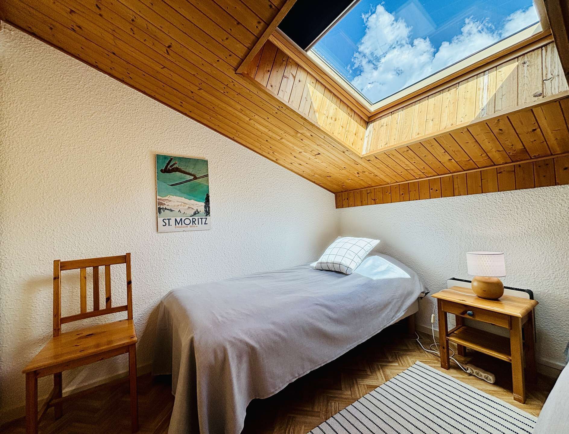 Un dormitorio pequeño con techo inclinado, una cama, una silla, una mesita y un cartel en la pared.