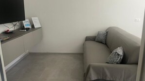 Una stanza con un divano grigio, una televisione a schermo piatto montata a parete, un tavolino con un telecomando e un cartello appeso al muro.