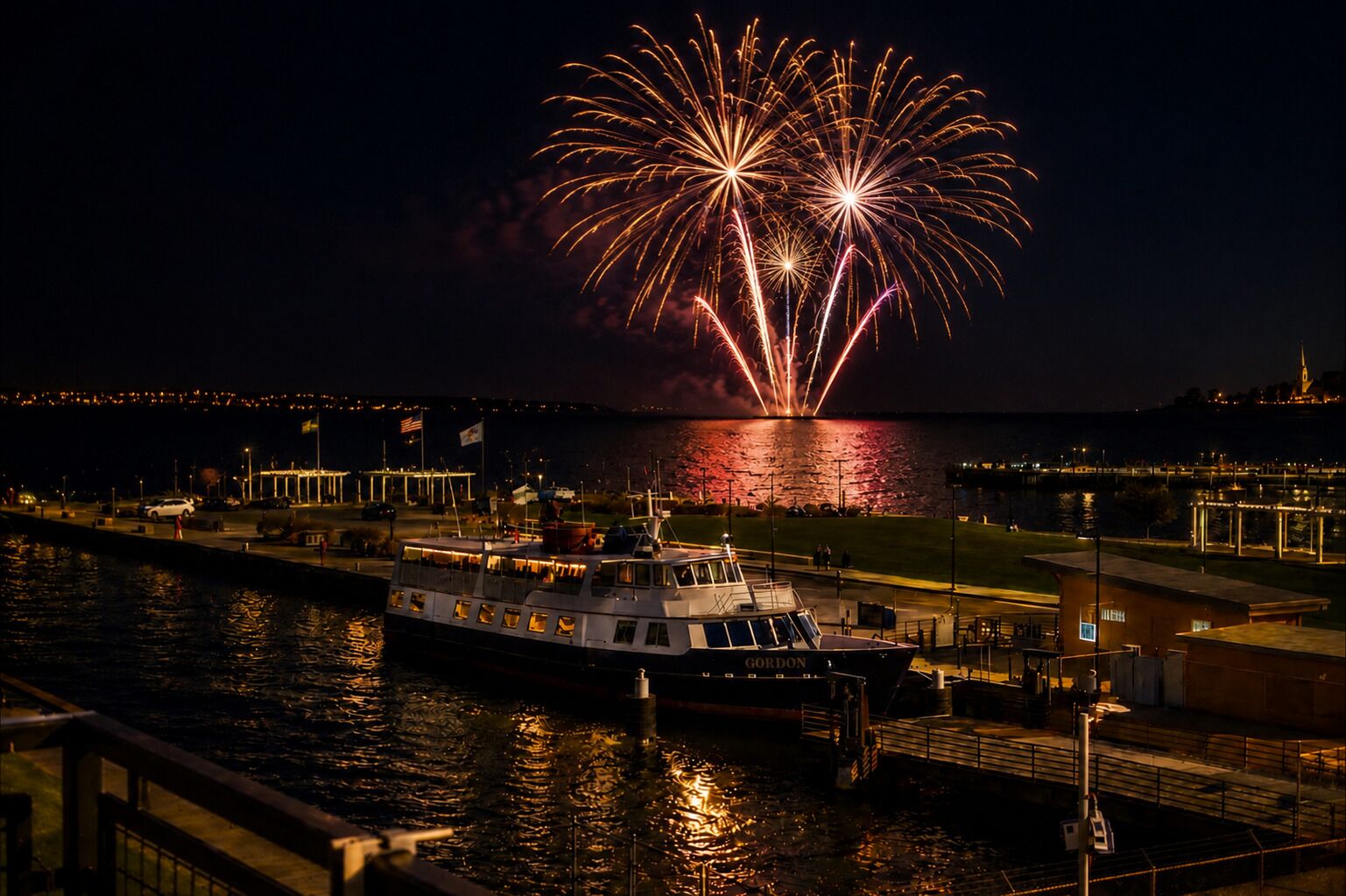 Une jetée au bord de l’eau où est amarré un bateau appelé « GORDON », un port de plaisance avec plusieurs bateaux, et un spectacle de feux d’artifice au-dessus de l’eau.