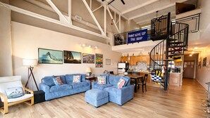 A spacious living area with a blue sofa set, a dining table, and a kitchen in the background. A sign reads 'DON'T GIVE UP THE SHIP'.