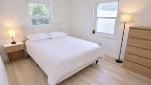 Ein Schlafzimmer mit einem Bett, Nachttischen, einer Kommode und zwei Fenstern.