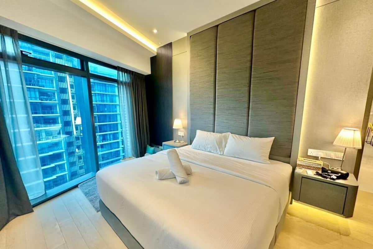 Habitación de hotel moderna con una cama grande, una mesita de noche, una lámpara y vistas a un paisaje urbano a través de ventanas amplias.