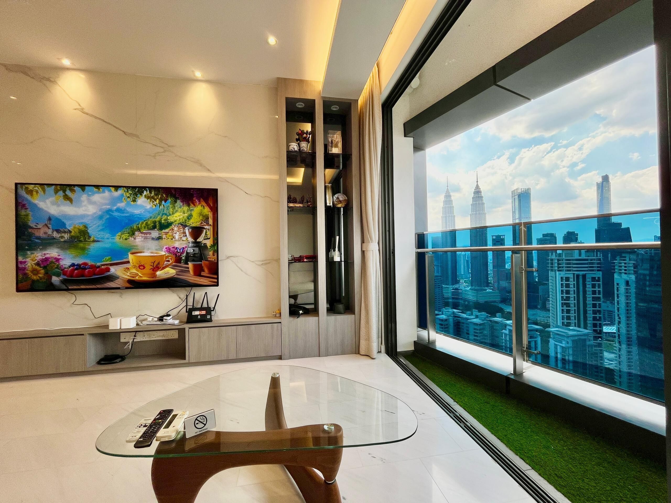 A modern living room with a glass table, a painting of a scenic landscape, a cityscape view through large windows, and a balcony with artificial grass.