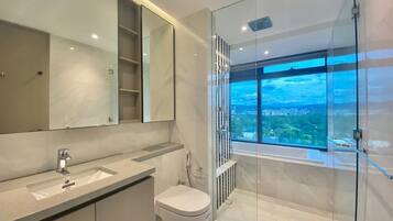Un baño moderno con un espejo grande, un inodoro, cabina de ducha y vistas a un paisaje urbano a través de un ventanal.