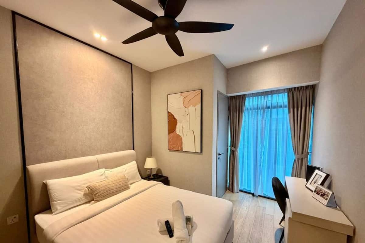Una habitación de hotel con cama, escritorio, silla, ventilador de techo, ventana con cortinas y un cuadro en la pared.