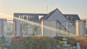 Een modern gebouw met een glazen gevel, een metalen dak en een tuin met struiken en een decoratieve fontein.