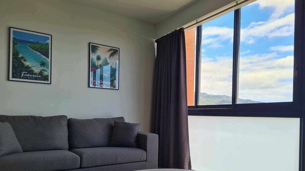 Een moderne woonkamer met een grijze bank, een ronde witte tafel en twee wanddecoraties met afbeeldingen van tropische landschappen.