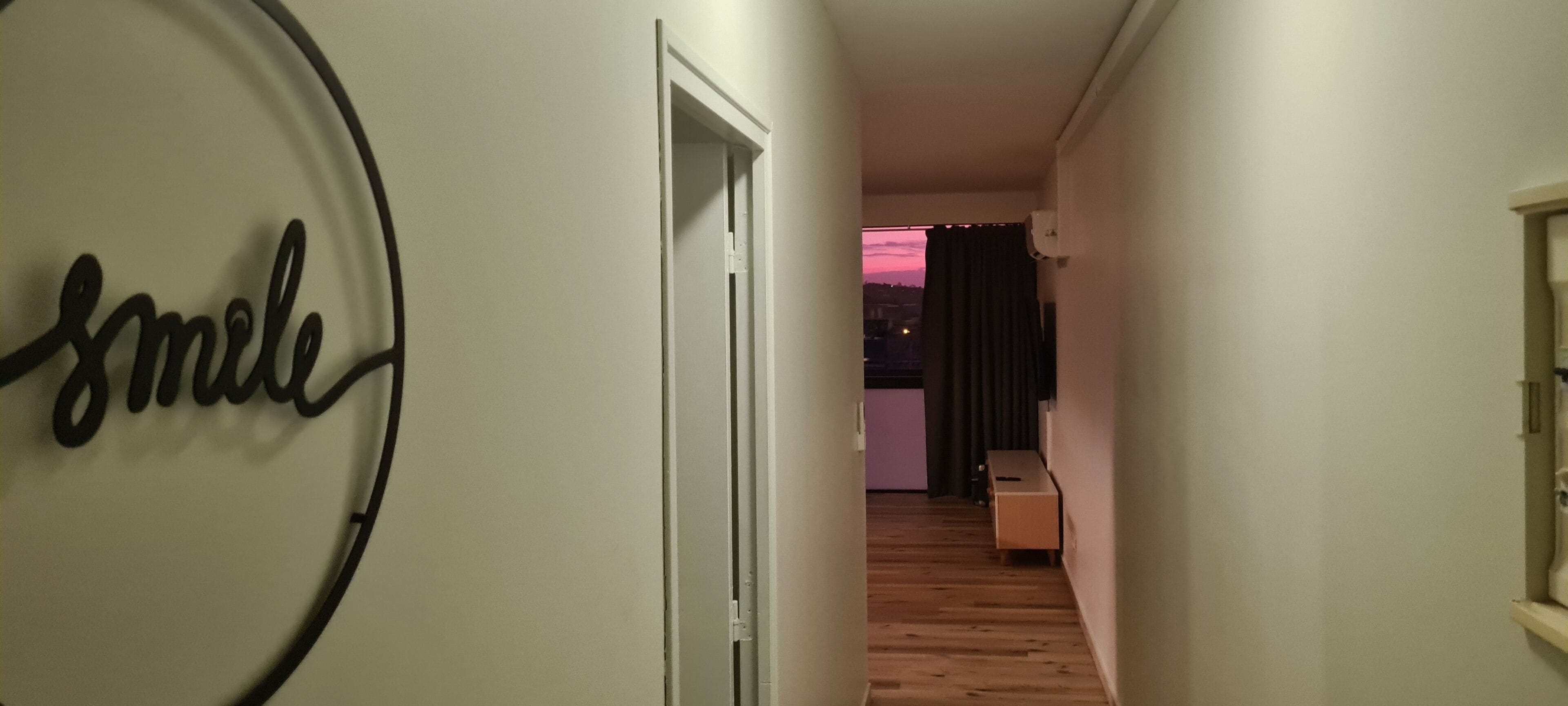 A hallway with a 'smile' sign, wooden flooring, a door leading to a room with a purple wall, and a window with curtains.