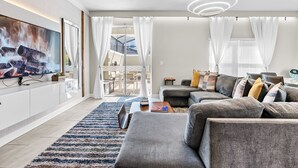 Ein modernes Wohnzimmer mit Flachbildfernseher, grauen Sofas, einem Couchtisch und einem großen Fenster mit Vorhängen.