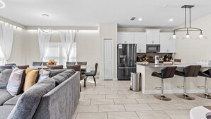 Ein modernes Wohnzimmer mit grauem Sofa, ein Essbereich mit Tisch und Stühlen, eine Küche mit weißen Schränken und Edelstahlgeräten sowie eine Bar mit Hockern.