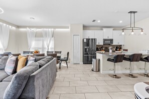 Ein modernes Wohnzimmer mit grauem Sofa, ein Essbereich mit Tisch und Stühlen, eine Küche mit weißen Schränken und Edelstahlgeräten sowie eine Bar mit Hockern.