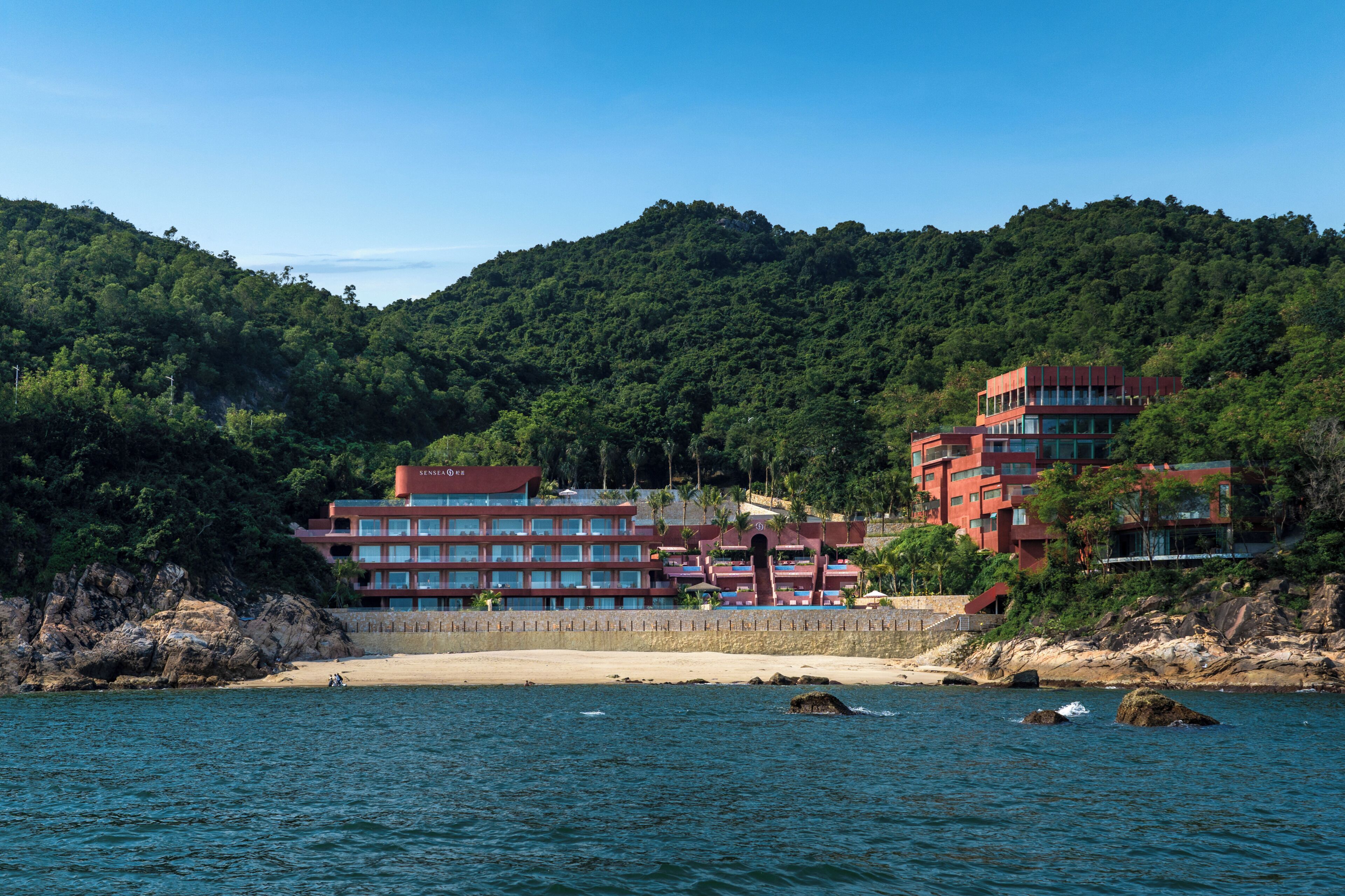 A coastal hotel with multiple floors, a beach in the foreground, and a forested hill in the background.