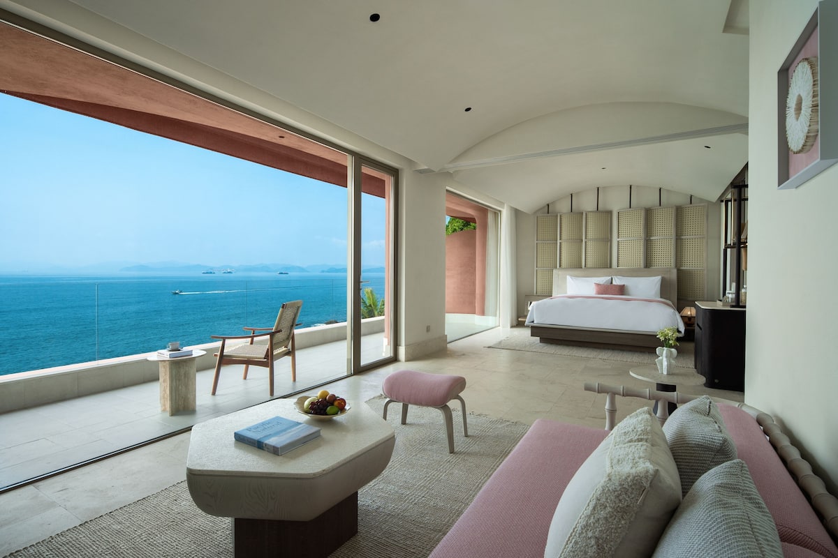 Un dormitorio moderno con una cama grande, una zona de estar con una mesa y un balcón con vistas al mar.