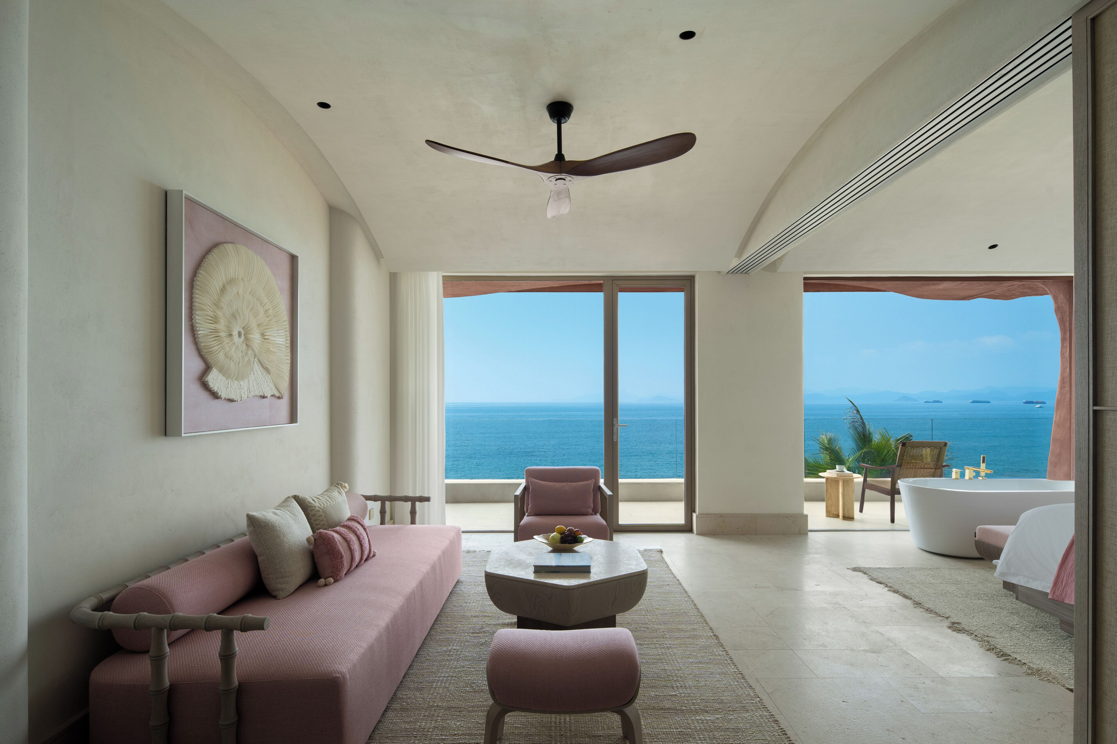 Una habitación moderna con un ventanal grande, ventilador de techo, sofá, mesa de centro y vista al océano.