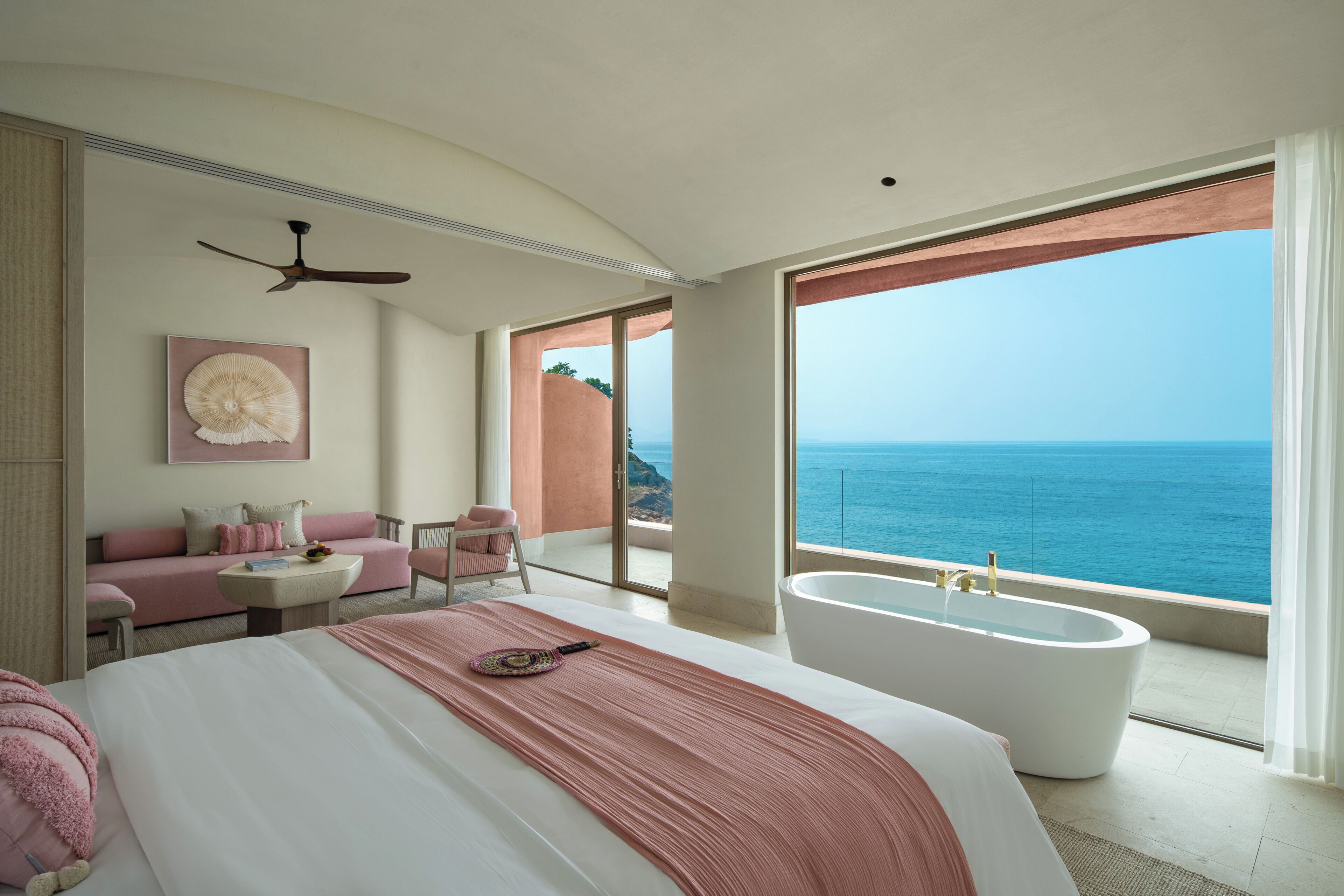Een slaapkamer met een groot bed, een badkuip, een plafondventilator, een sofa en uitzicht op de oceaan.