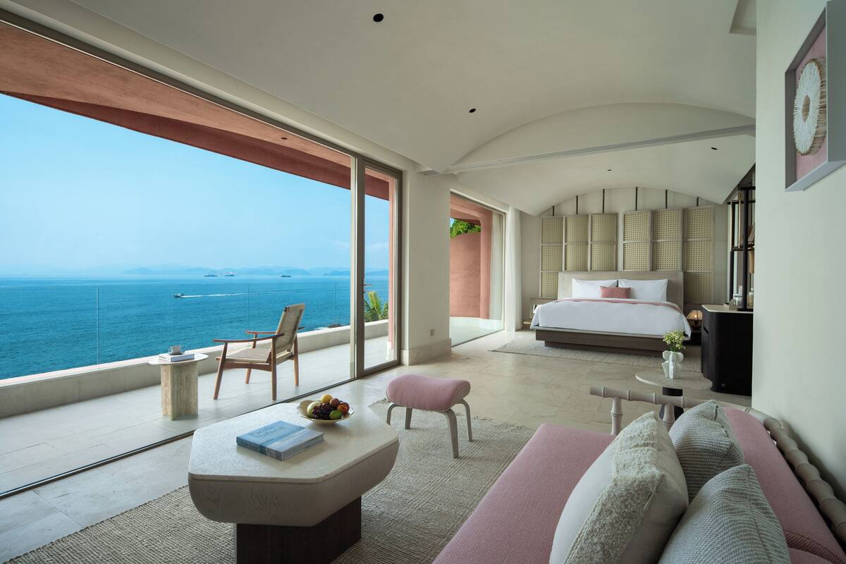 Una habitación amplia con una cama grande, un área de descanso con una mesa y un balcón con vista al mar.
