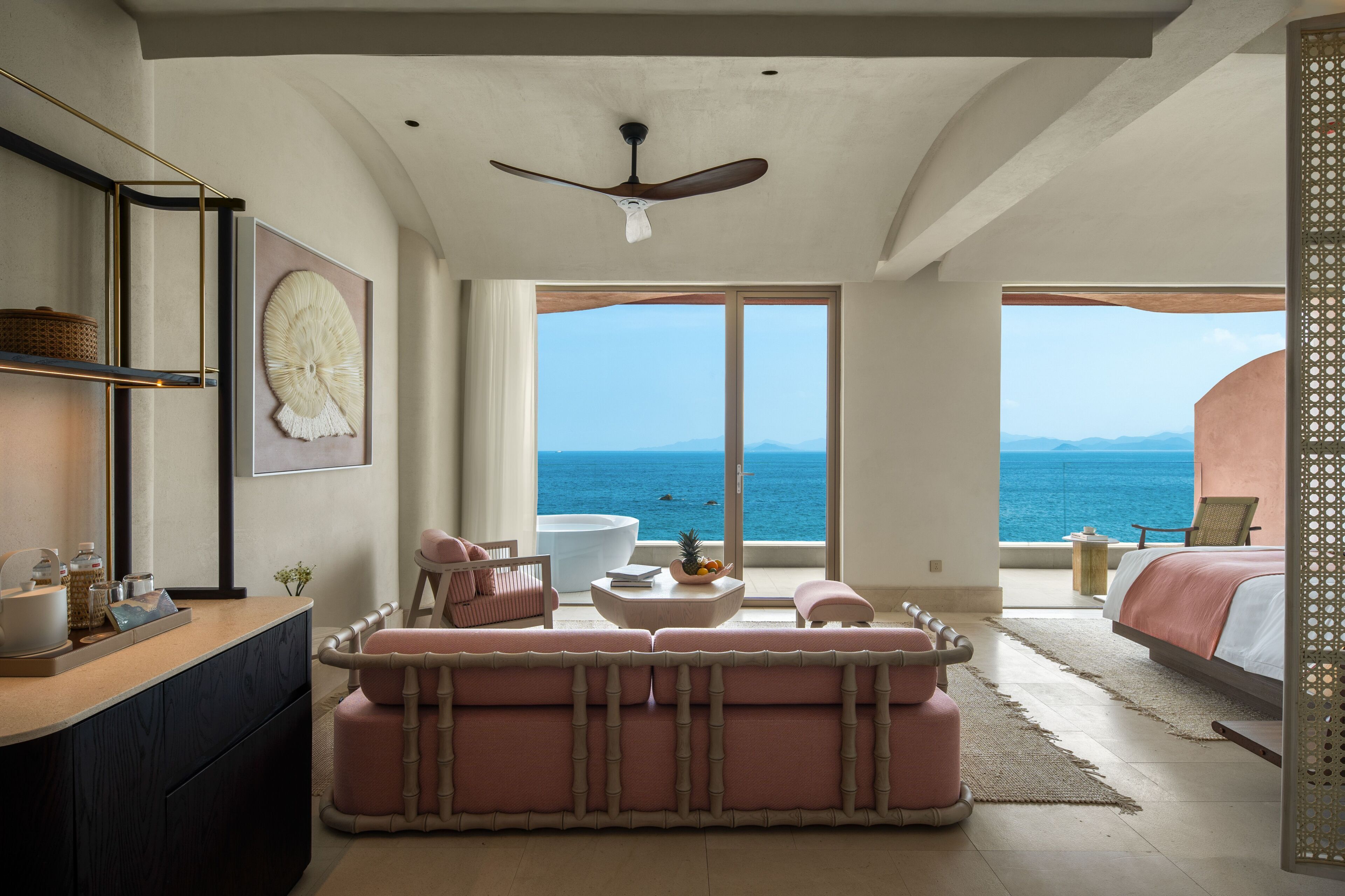 Una habitación amplia con una cama grande, un área de estar con un sofá y sillones, una mesita con un cuenco de fruta, y vista al océano a través de ventanas amplias.