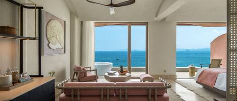 Una habitación amplia con una cama grande, un área de estar con un sofá y sillones, una mesita con un cuenco de fruta, y vista al océano a través de ventanas amplias.