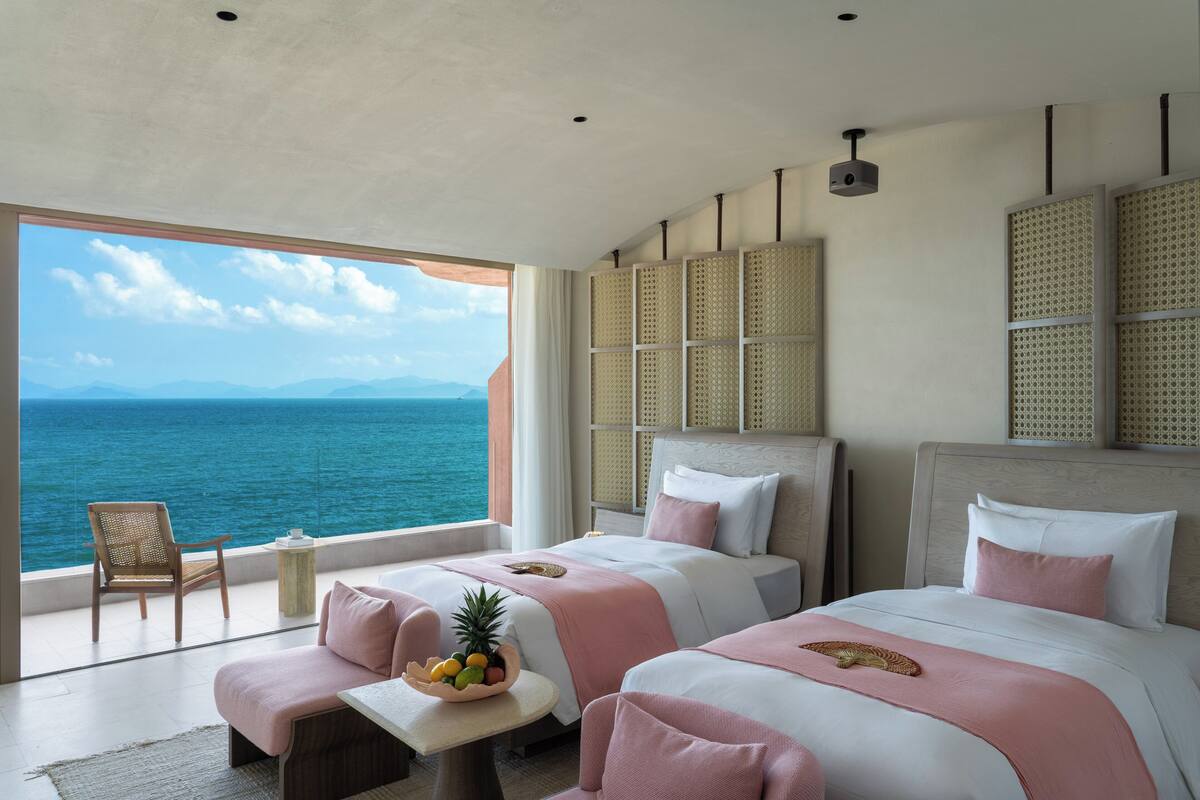 Habitación de hotel con dos camas, vista al mar, cabecero de mimbre y una pequeña mesa con fruta.