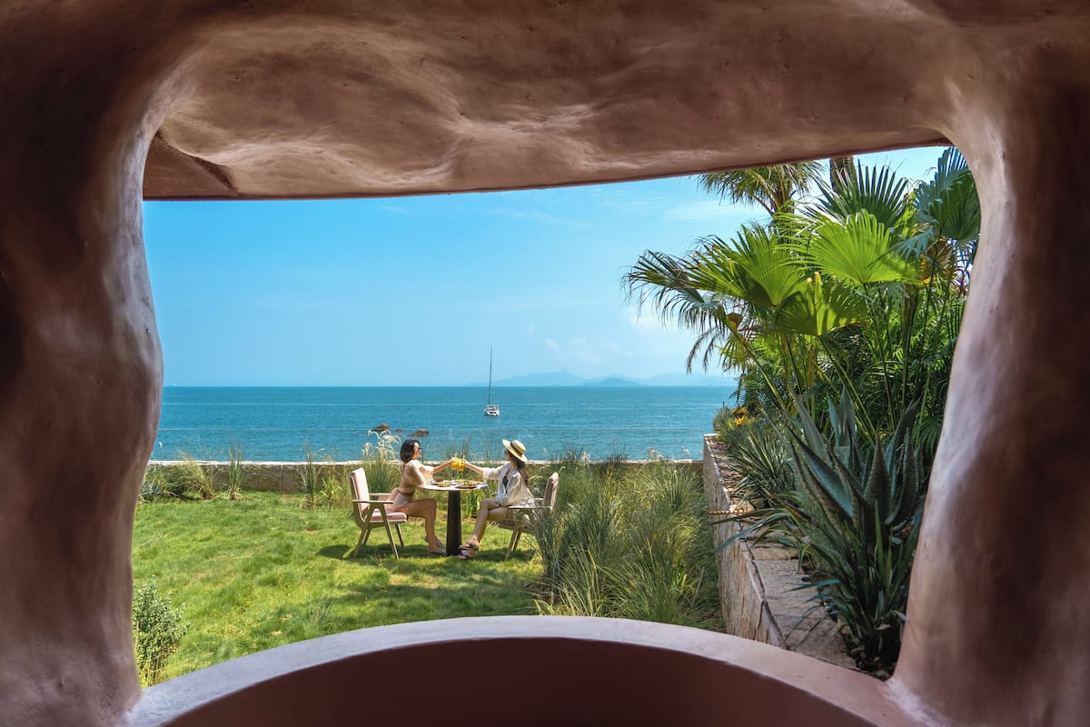 Vista desde una habitación con un arco de piedra que da a un espacio verde con una mesa y sillas, un cuerpo de agua y palmeras.