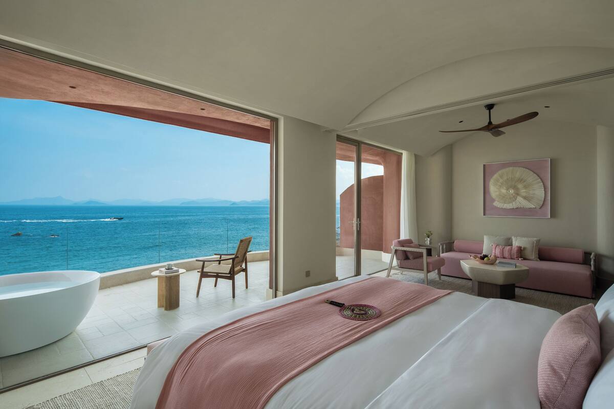 Un dormitorio con una cama grande, vista al mar, ventilador de techo y zona de estar con una mesa y sillas.
