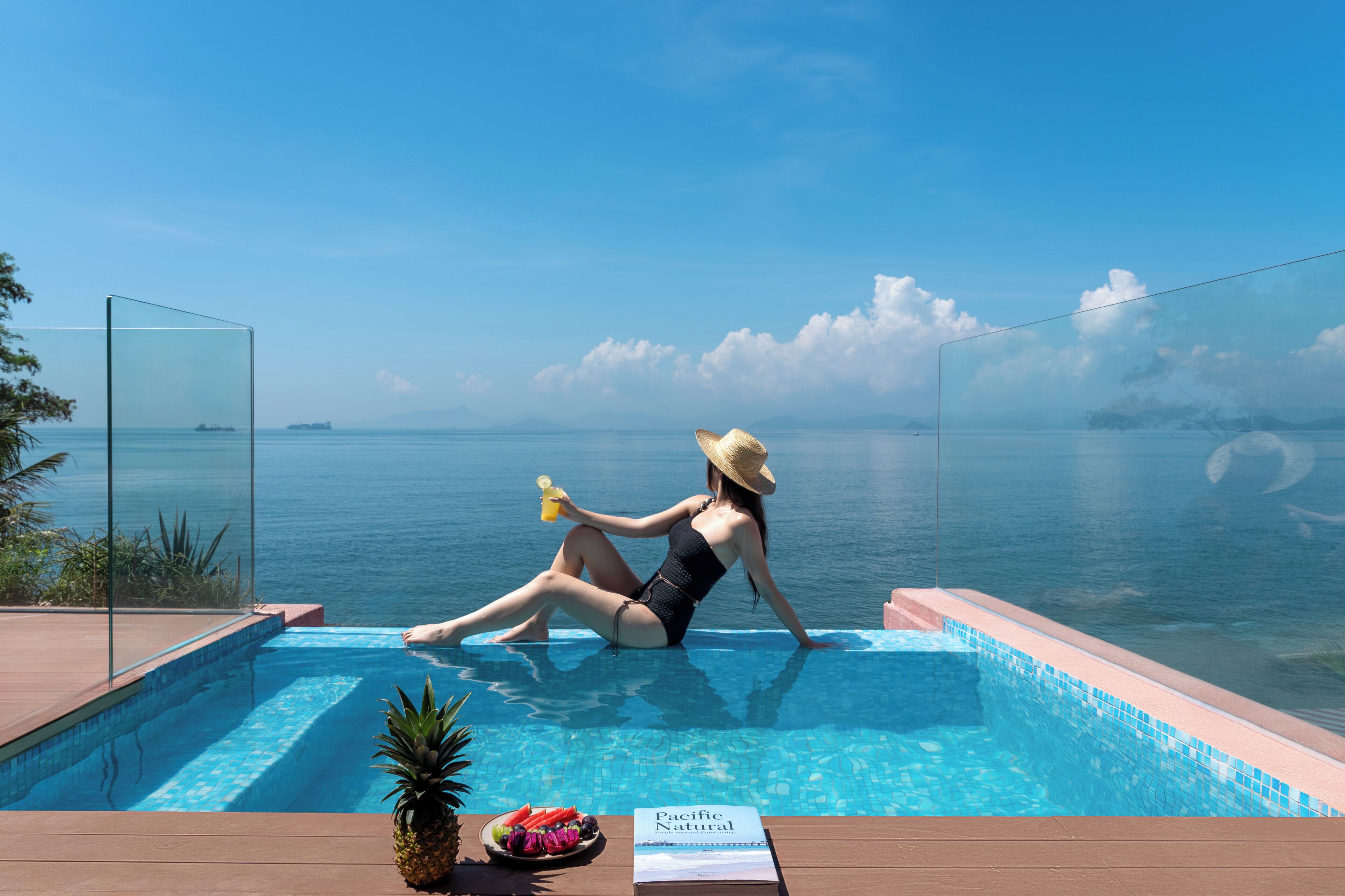 A woman in a swimsuit sitting by a pool with a view of the sea, a pineapple, and a book titled 'Pacific Natural' on the deck.