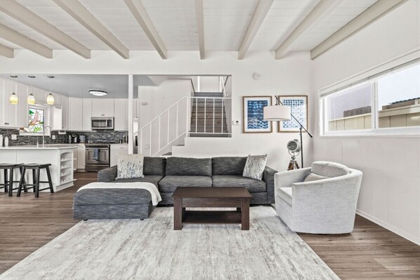 A modern living room with a sofa, ottoman, and armchair, a coffee table, and a staircase leading to an upper level. - South Mission Beach Home – 3BR, Epic Patio, Rare 3-Car Parking, Steps to the Sand (San Diego)