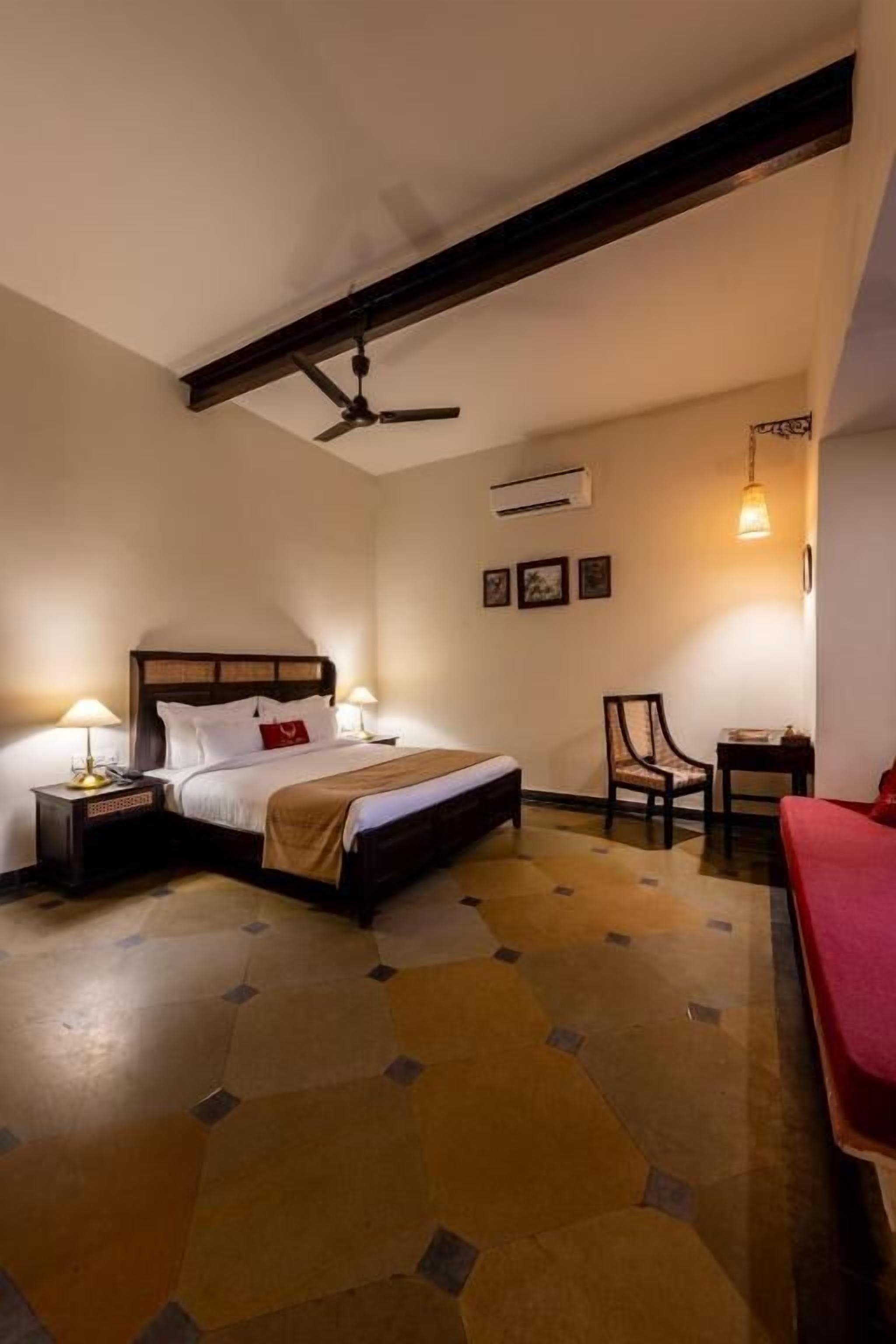 A hotel room with a bed, two bedside tables, a chair, a red sofa, a ceiling fan, and a wall-mounted air conditioner.