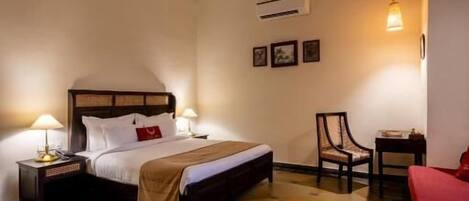 A hotel room with a bed, two bedside tables, a chair, a red sofa, a ceiling fan, and a wall-mounted air conditioner.